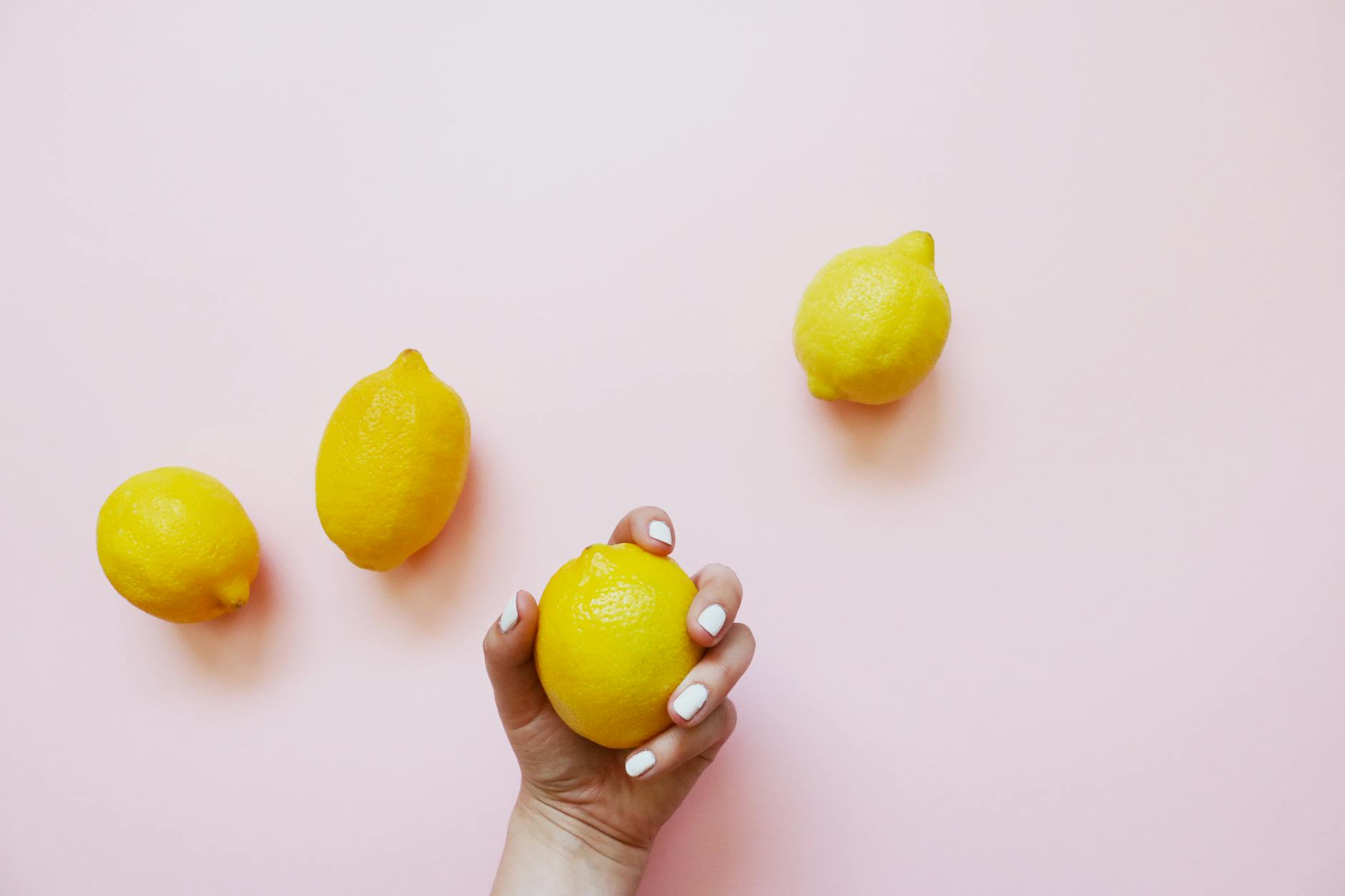 A hand with white nails holding a fresh lemon on a soft pink background, symbolizing the bright, citrusy sensation of exploring pleasure solo.